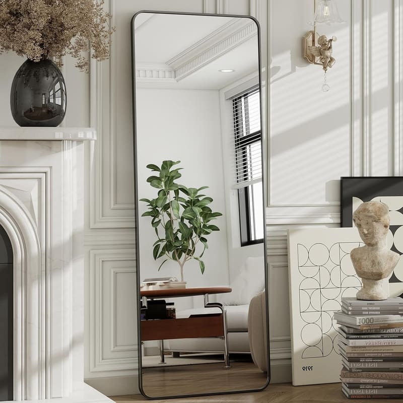 The Artment Vista mirror lifestyle shot in a Pinterest-style bedroom corner with a houseplant and a small wooden stool, showing the mirror leaning at the recommended 5-10° tilt against the wall