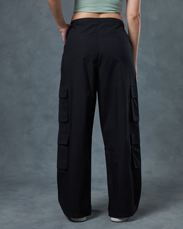 Full-length styling shot of the Bewakoof Jet Black parachute pants worn with a sage ribbed tank top and white sneakers